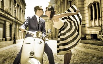 From Italy To Bossa With Love. A well-dressed man kisses a flambouyant women wearing a stunning black and white sun dress hile sitting on a Vespa motorbike on an Italian cobblestone street.