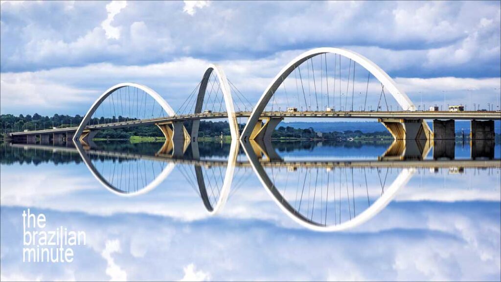The arches of the Juscelino Kubitschek Bridge are reflected over water on a cloud-touched day.