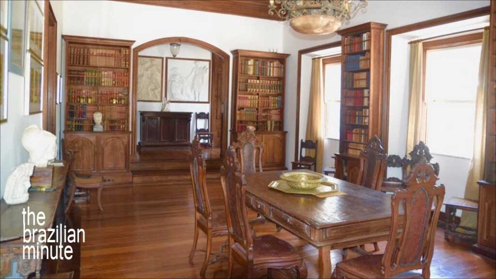 Rio de Janeiro's Historic Villa Riso. The library room includes an ornate wood table with six matching chaird plus four tall wooden bookcases and two windows with open curtains.