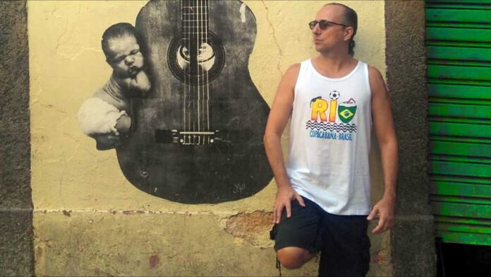 Brazilian guitarist Phill Fest Sings a Slice of Life, standing in front of a yelllow wall featurng a street art guitar.