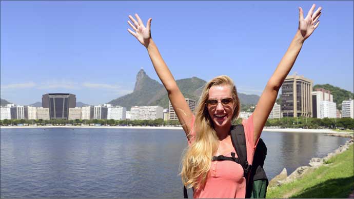 Thin young blonde woman with outstretched arms smiling in front of Rio de Janeiro's Lagoa.
